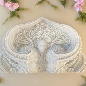 May include: A white ceramic wall sculpture with intricate details and a face in the center. The sculpture is shaped like a heart and has a textured surface.
