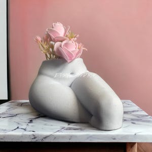May include: A white ceramic planter shaped like a pair of legs with pink flowers inside. The planter is sitting on a marble surface.