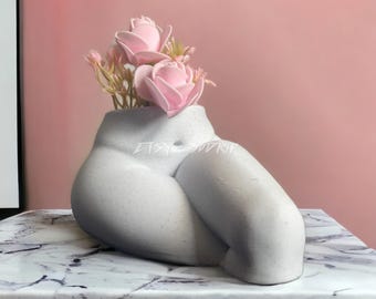 May include: A white ceramic planter shaped like a pair of legs with pink flowers inside. The planter is sitting on a marble surface.