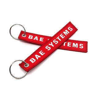 Könnte beinhalten: Zwei rote Schlüsselanhänger mit weiß gesticktem Text. Der obere Schlüsselanhänger trägt die Aufschrift "BAE SYSTEMS". Der untere Schlüsselanhänger trägt die Aufschrift "SYSTEMS".