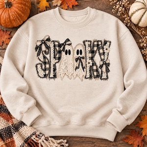 Puede incluir: Una sudadera de cuello redondo en color beige con la palabra SPOOKY en una fuente decorativa. Las letras están rellenas con un patrón de cuadros en blanco y negro, adornadas con encaje blanco, lazos de tela y dos figuras de fantasmas.