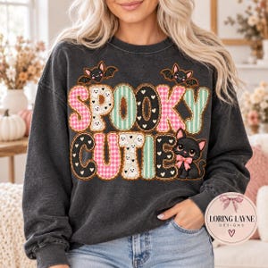 Peut inclure: Un sweat-shirt gris anthracite à col rond avec le texte SPOOKY CUTIE dans une police à motifs variés. Les lettres sont décorées de fantômes, de vichy rose et de rayures bleu sarcelle. Deux petites chauves-souris et un chaton noir avec un nœud rose.