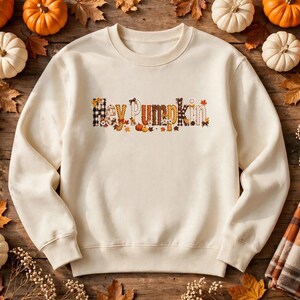 Op de afbeelding: Een crèmekleurige sweater met ronde hals en de tekst Hey Pumpkin op de borst gedrukt. De letters zijn versierd met diverse herfstthema-patronen, waaronder zwart-wit geblokte ruiten, oranje bladeren en kleine pompoenen.
