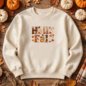 May include: A cream-colored crewneck sweatshirt featuring the text "Hello Fall" printed on the chest. The letters are decorated with various autumn-themed patterns including plaid, polka dots, and small illustrations of pumpkins and leaves.
