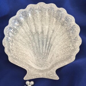 May include: A translucent, shell-shaped dish featuring a scalloped edge and a textured surface embedded with fine silver glitter particles. The dish has a concave centre and a wide, fan-like form, reflecting light across its shimmering, clear finish.