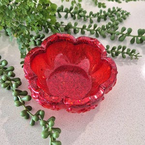 May include: A vibrant red, flower-shaped bowl featuring a translucent finish embedded with fine red glitter throughout the material. The bowl has five rounded petals forming the rim and a smooth, circular base, creating a decorative vessel.