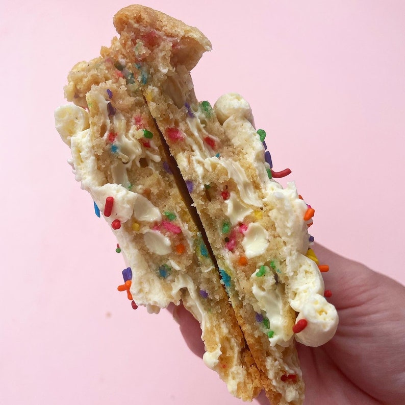 K&ouml;nnte beinhalten: Nahaufnahme eines geteilten Geburtstags-Kekses. Der Keks ist mit wei&szlig;er Zuckerglasur und bunten Streuseln gef&uuml;llt. Der Keks wird in einer Hand gehalten.