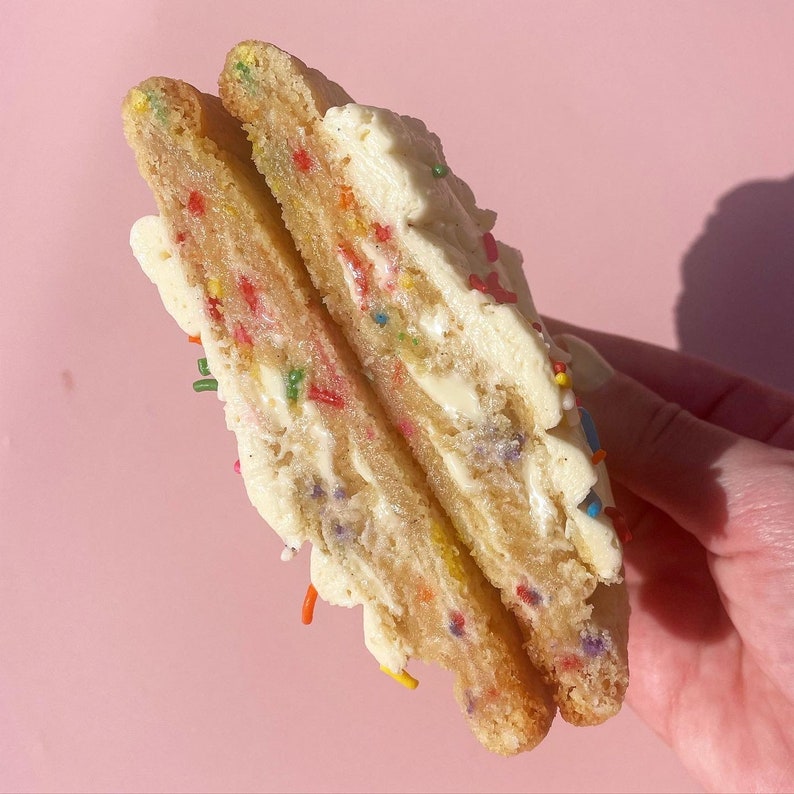 K&ouml;nnte beinhalten: Nahaufnahme eines geteilten Keks-Sandwiches mit Vanille-Frosting und bunten Streuseln. Der Keks ist hellbraun und das Frosting ist wei&szlig;.