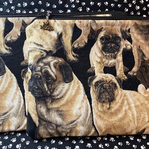 May include: Two zippered pouches with a repeating pattern of pug dogs in various poses on a black background. The pugs are tan and black. The zippers have decorative charms. The background fabric has a white paw print pattern.