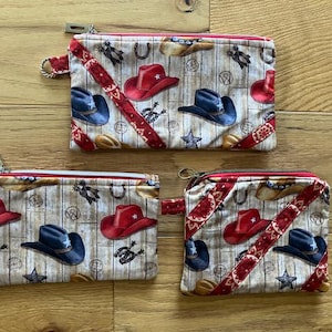 May include: Three zippered pouches with a cowboy theme. The pouches are made of fabric with a beige background and a red, white, and blue pattern of cowboy hats, horseshoes, and stars. The pouches are different sizes.