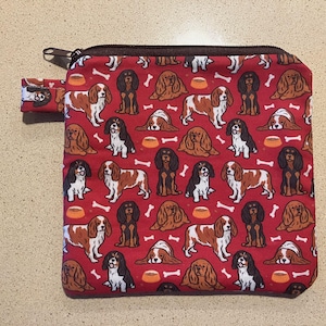 以下が含まれることがあります： キャバリア・キング・チャールズ・スパニエルの犬、犬のボウル、骨の繰り返し模様が施された赤いジッパーポーチ。ポーチには茶色のジッパーと小さな布ループが付いています。