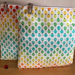 May include: Two white zippered pouches with a repeating pattern of colorful ladybugs on a white background. The pouches are lined up side by side on a wooden surface.