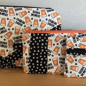 May include: Three zippered pouches with a white background and black paw prints. The pouches have a repeating pattern of orange beer mugs with the text "Drinking Buddy".