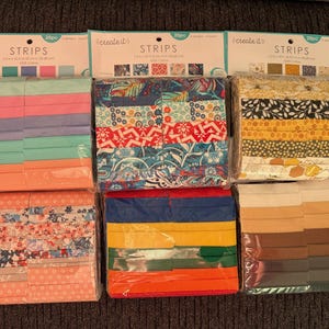 May include: Six packs of fabric strips in assorted colours and patterns. Each pack has a label reading "create it" and "STRIPS". The fabric strips feature shades of pink, blue, orange, red, yellow, and brown, with floral and geometric designs.