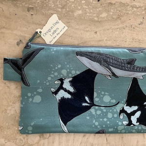 May include: A teal zippered pouch featuring illustrations of marine life, including a whale shark, manta rays, and a whale tail. The pouch has a small fabric loop and a tag that reads "Oregon Bag Ladies."