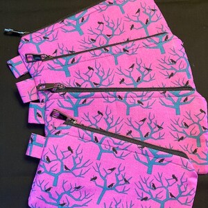 Könnte beinhalten: Eine Sammlung pinkfarbener Reißverschlusstaschen mit einem sich wiederholenden Muster aus türkisfarbenen Bäumen und schwarzen Vögeln. Jede Tasche hat einen schwarzen Reißverschluss und eine kleine Stoffschlaufe. Auf schwarzem Untergrund.