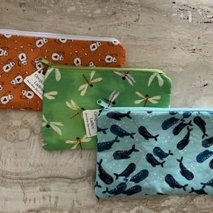 May include: Three zippered pouches with unique patterns. One is orange with cartoon figures, another is green with dragonflies, and the last is light blue with narwhals. Each pouch has a white tag with text.