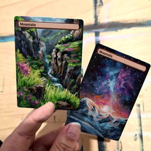 Puede incluir: Dos cartas de "Montaña" pintadas a mano. Una representa un arroyo de montaña con hierba verde y flores rosas. La otra muestra un cielo estrellado sobre una montaña nevada. Las cartas tienen un borde marrón y la palabra "Montaña" en la parte superior.