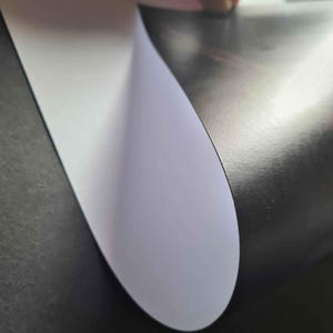 May include: A close-up of a white sheet of paper being peeled back from a black surface. The paper is slightly curved and has a smooth, matte finish.