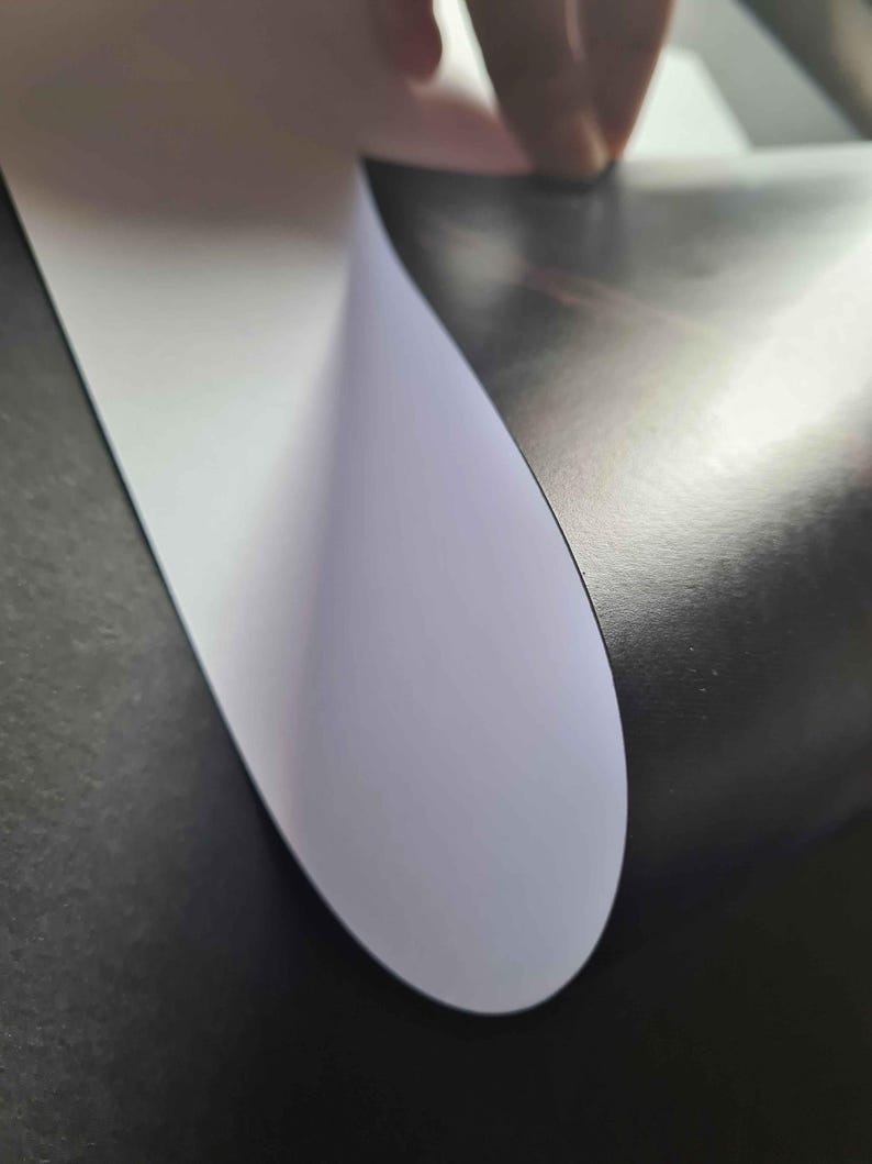 May include: A close-up of a white sheet of paper being peeled back from a black surface. The paper is slightly curved and has a smooth, matte finish.