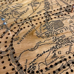 May include: Wooden cribbage board featuring a detailed map of Middle-earth, with place names like Shire and Rohan. The board has numerous holes for pegs and is made of light-colored wood. Two pegs are visible, one silver and one copper.