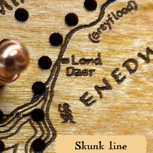 May include: Close-up of a wooden map with engraved details and dark circular accents. The text "Land Deer" and "Skunk line" are visible. A copper-colored knob is in the upper left corner.