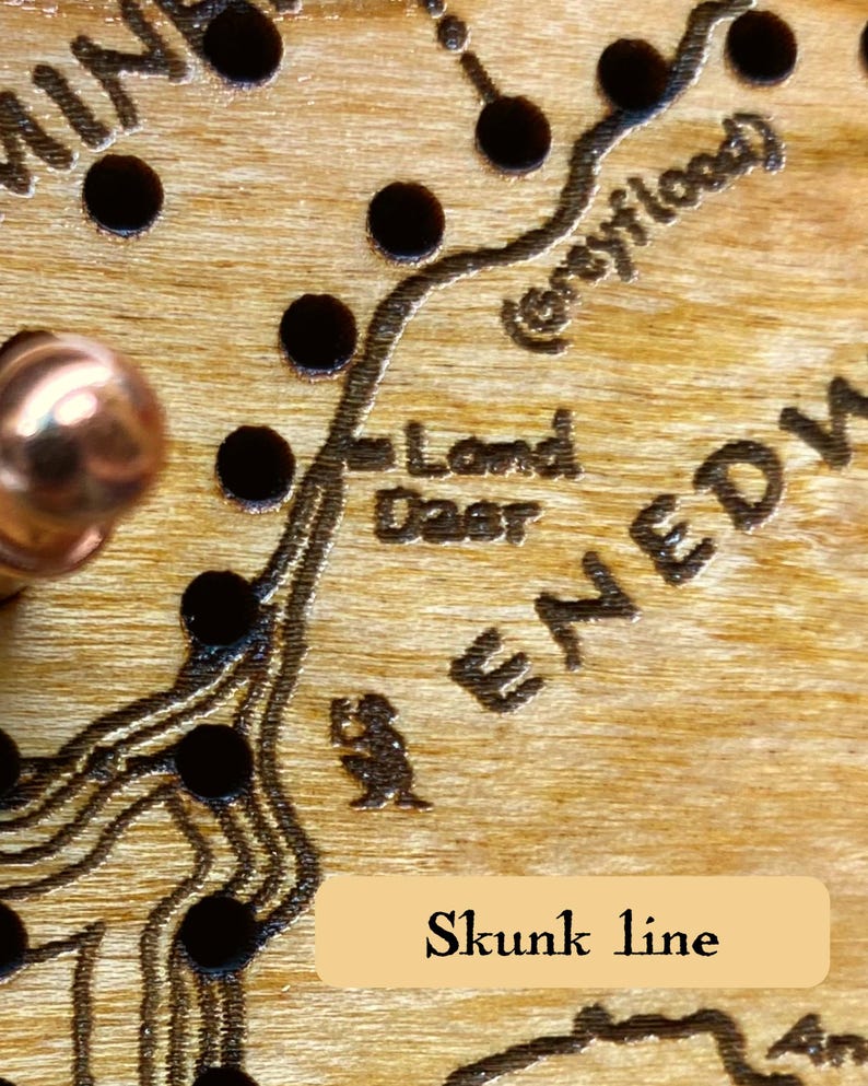 May include: Close-up of a wooden map with engraved details and dark circular accents. The text "Land Deer" and "Skunk line" are visible. A copper-colored knob is in the upper left corner.