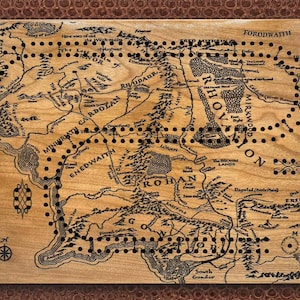 May include: Wooden cribbage board featuring a detailed map of Middle-earth with black peg holes. The board has a light brown finish with dark brown text and illustrations, including place names like Mordor and Rohan. The board is rectangular and has a dark brown border.