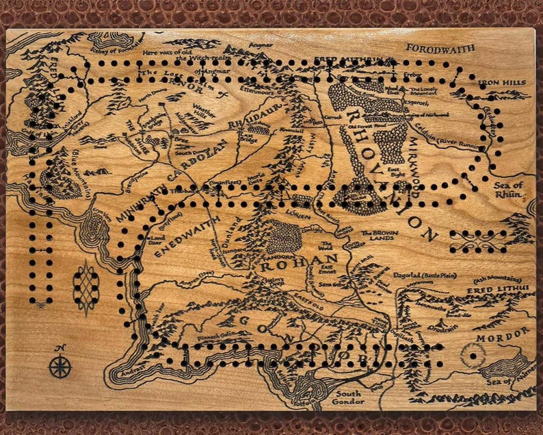 May include: Wooden cribbage board featuring a detailed map of Middle-earth with black peg holes. The board has a light brown finish with dark brown text and illustrations, including place names like Mordor and Rohan. The board is rectangular and has a dark brown border.