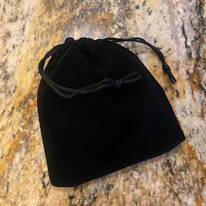 May include: A black velvet drawstring bag with a black cord closure. The bag is closed and sits on a speckled, tan and brown surface. The bag is likely used for storing jewelry or small items.