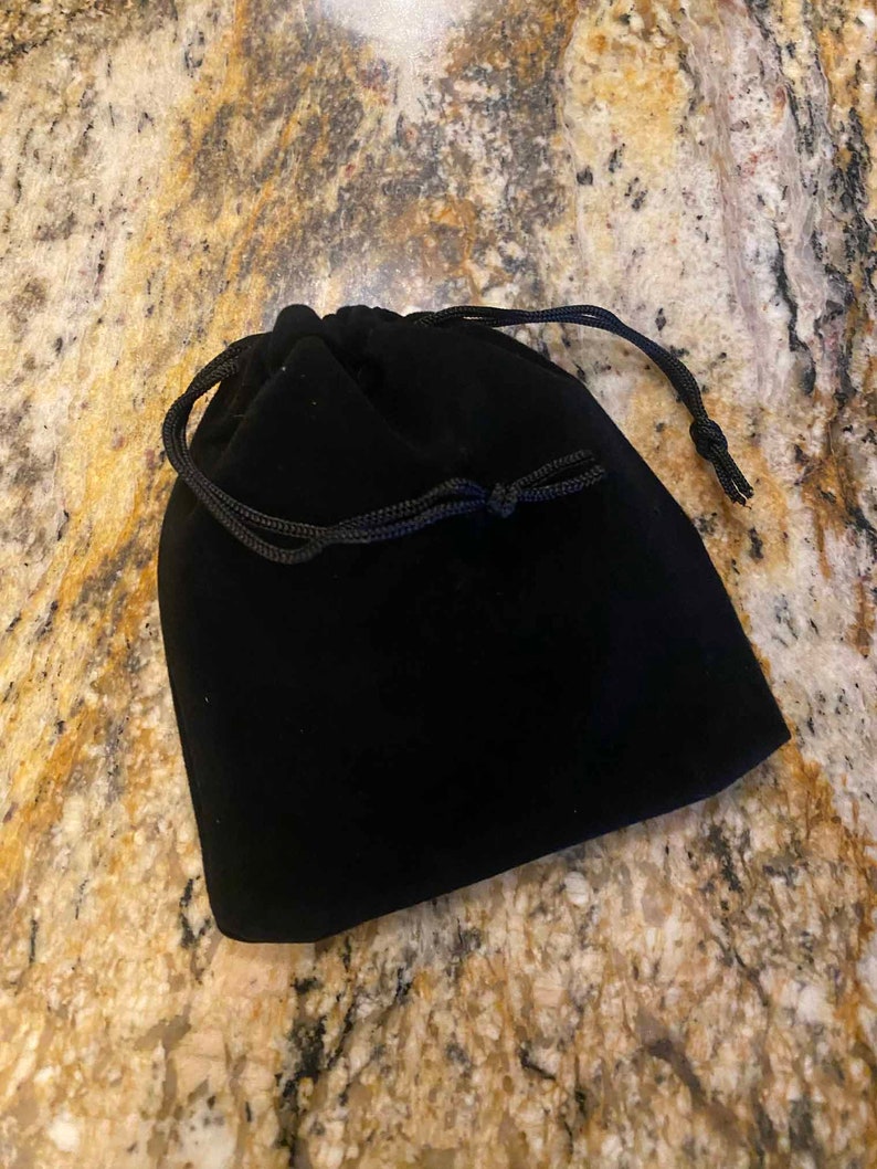 May include: A black velvet drawstring bag with a black cord closure. The bag is closed and sits on a speckled, tan and brown surface. The bag is likely used for storing jewelry or small items.