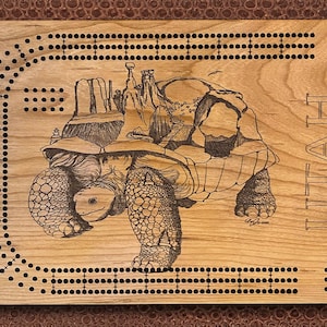 Può includere: Un tabellone di cribbage in legno con un design inciso a laser di una tartaruga che trasporta un paesaggio con il testo "UTAH Gateway to the Parks".