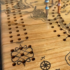 May include: Close-up of a wooden cribbage board with a map design and decorative elements. The board has a light wood grain, with engraved lines and holes for pegs. Three metal pegs are inserted. The text "The Realm Of" and a compass rose are visible.
