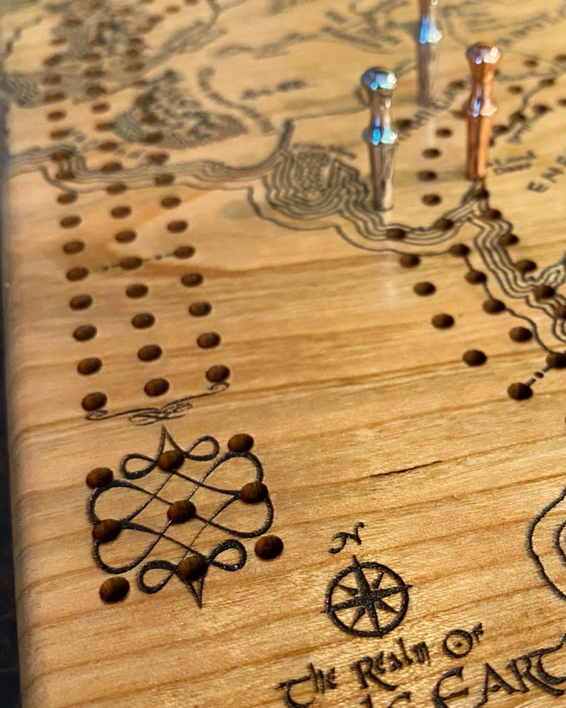 May include: Close-up of a wooden cribbage board with a map design and decorative elements. The board has a light wood grain, with engraved lines and holes for pegs. Three metal pegs are inserted. The text "The Realm Of" and a compass rose are visible.