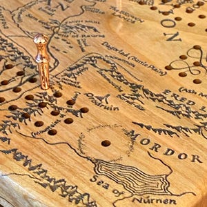 May include: Wooden cribbage board featuring a detailed map of Middle-earth, with place names like Mordor and Rohan. The board has a light wood grain and includes copper-colored pegs. The map is etched in black.