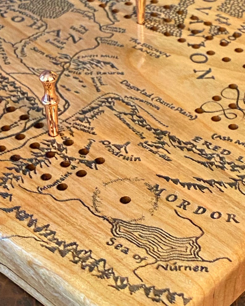 May include: Wooden cribbage board featuring a detailed map of Middle-earth, with place names like Mordor and Rohan. The board has a light wood grain and includes copper-colored pegs. The map is etched in black.