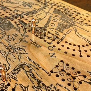 May include: Wooden cribbage board with a map design, featuring etched details and peg holes. The board has a light wood grain, with copper-colored pegs inserted. The map includes text such as "Mirkwood" and "Ered Lith".