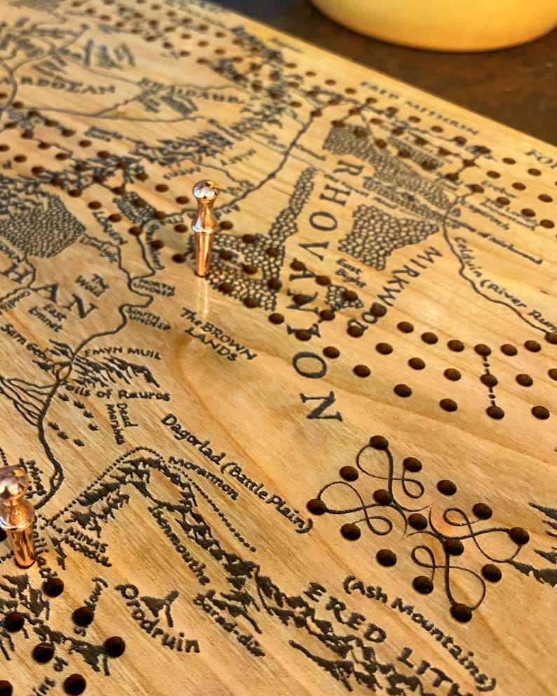 May include: Wooden cribbage board with a map design, featuring etched details and peg holes. The board has a light wood grain, with copper-colored pegs inserted. The map includes text such as "Mirkwood" and "Ered Lith".