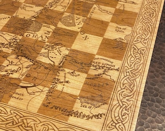 May include: A square wooden chessboard featuring a laser-engraved map of Middle-earth. The board has alternating light and dark wood squares, with intricate Celtic knotwork borders and a tree symbol in the corner. Place names like Rohan, Mordor, and Mirkwood are clearly visible across the surface.