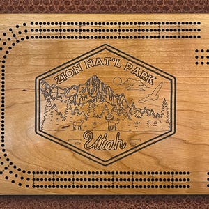 May include: A wooden cribbage board with a laser-engraved design featuring a mountain scene and the text "Zion Nat'l Park - Utah".