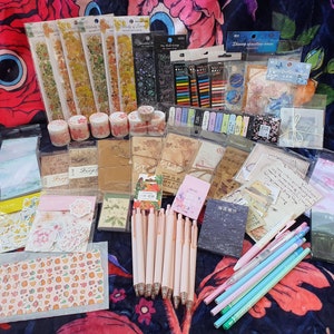 May include: A collection of craft supplies including washi tape, decorative paper, stickers, pens, and other stationery items. The items are arranged on a floral patterned fabric.