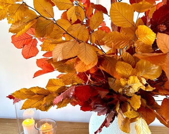May include: A white textured ceramic vase filled with vibrant autumn beech branches featuring orange, red, and yellow leaves. Beside the vase are two clear glass cylindrical candle holders, each containing a small white pillar candle with a warm glow.