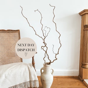 May include: A beige circular sign with the text "NEXT DAY DISPATCH" and a small heart symbol. The sign is leaning against a wicker chair with a white throw blanket. A vase with dried branches is in the foreground.