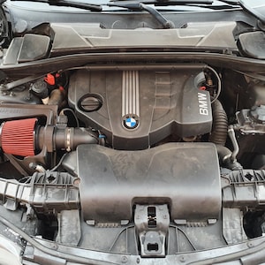 Könnte beinhalten: Nahaufnahme eines schwarzen BMW-Motors mit einem roten Luftfilter. Der Motor ist offen und die Motorhaube ist angehoben. Der Motor hat eine schwarze Kunststoffabdeckung mit dem BMW-Logo. Der Luftfilter ist ein rotes zylindrisches Objekt mit einem schwarzen Gehäuse. Der Motor ist sauber und gut gewartet.