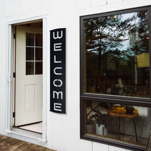 Pu&ograve; includere: Un cartello WELCOME in metallo nero &egrave; montato verticalmente su un muro esterno bianco accanto a una porta. Il cartello riporta la parola WELCOME in lettere maiuscole bianche. Sono visibili anche una finestra e un terrazzo in legno.
