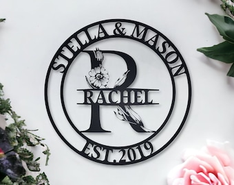 Puede incluir: Círculo de metal negro con el texto "Stella & Mason Est. 2019" alrededor del exterior. La letra "R" está en el centro con un diseño floral y el nombre "Rachel" escrito en el centro.