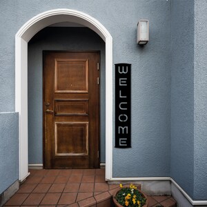 Pu&ograve; includere: Una porta in legno marrone scuro &egrave; incorniciata da un arco bianco. Un cartello verticale nero con la scritta "WELCOME" in lettere bianche &egrave; appeso alla parete blu. Un piccolo vaso di fiori con fiori gialli &egrave; vicino all'ingresso.