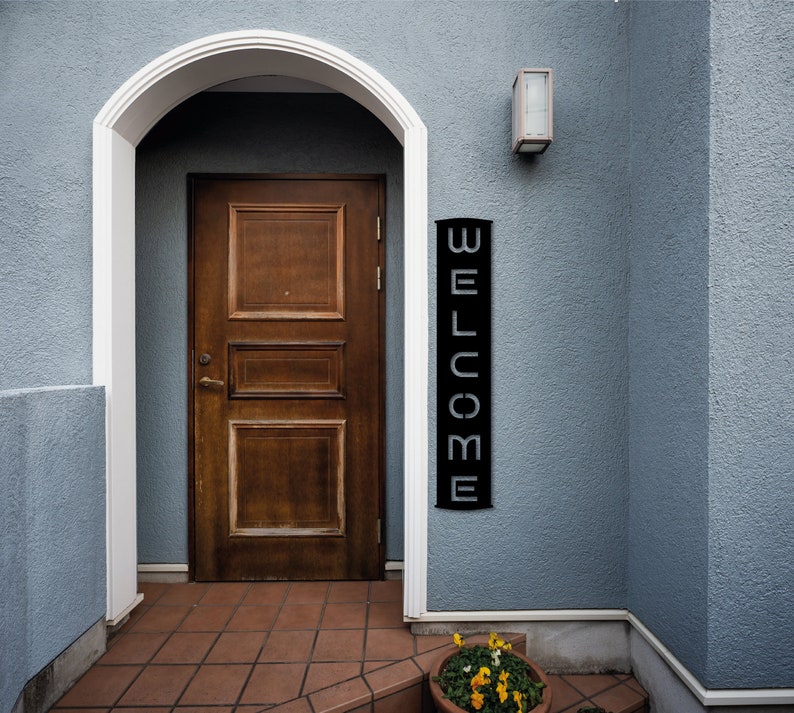 Pu&ograve; includere: Una porta in legno marrone scuro &egrave; incorniciata da un arco bianco. Un cartello verticale nero con la scritta "WELCOME" in lettere bianche &egrave; appeso alla parete blu. Un piccolo vaso di fiori con fiori gialli &egrave; vicino all'ingresso.