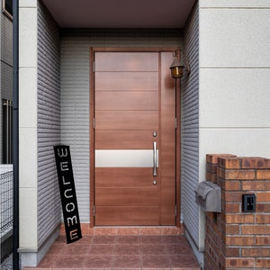 Pu&ograve; includere: Una porta d'ingresso in legno marrone con un pannello di vetro smerigliato orizzontale e una maniglia argentata. Un cartello nero con la scritta "WELCOME" &egrave; appoggiato sui gradini. Una cassetta delle lettere in mattoni e una lampada da parete vintage sono visibili.