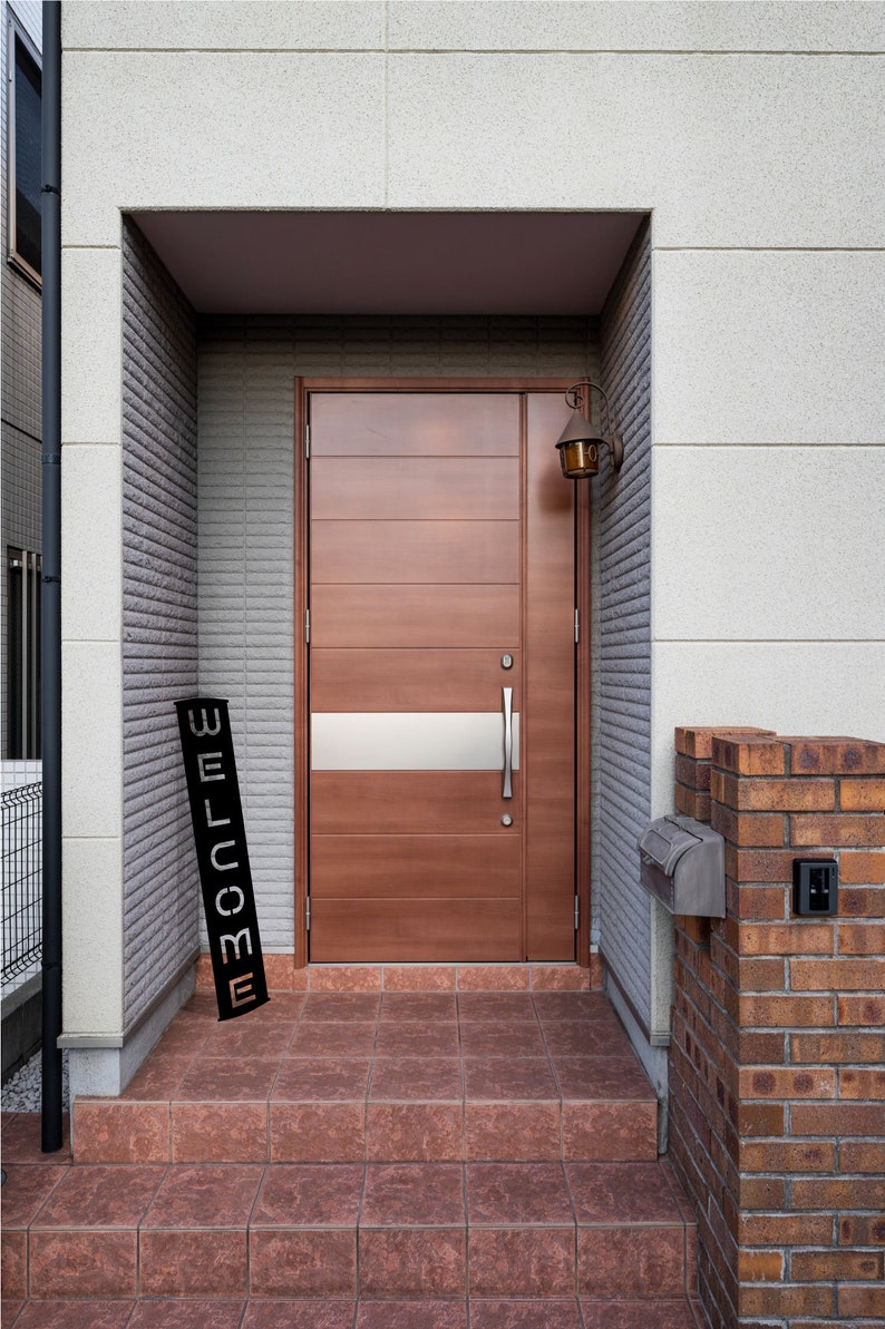 Pu&ograve; includere: Una porta d'ingresso in legno marrone con un pannello di vetro smerigliato orizzontale e una maniglia argentata. Un cartello nero con la scritta "WELCOME" &egrave; appoggiato sui gradini. Una cassetta delle lettere in mattoni e una lampada da parete vintage sono visibili.